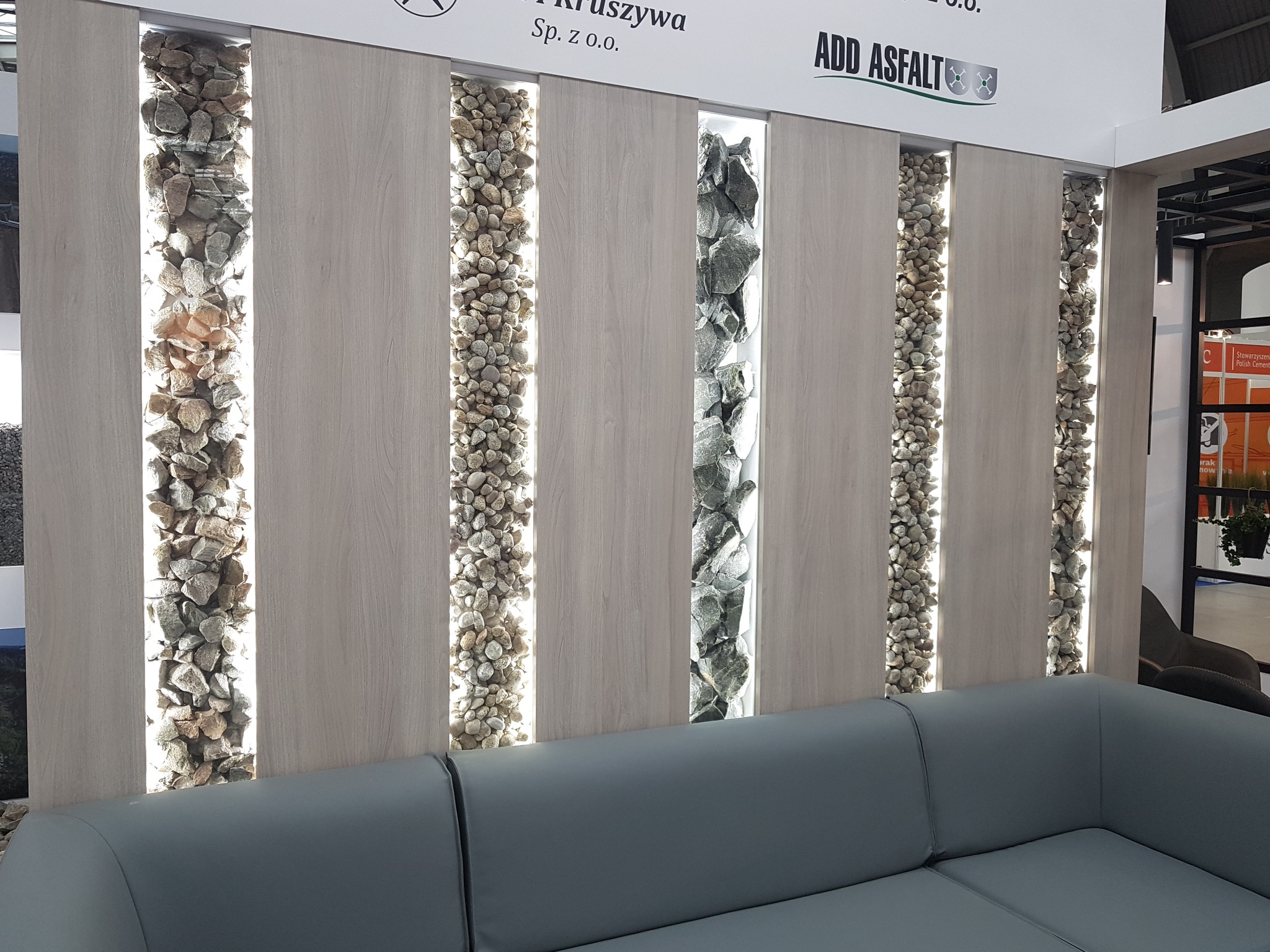 Panel dekoracyjny z podświetlanymi wnękami wypełnionymi kamieniami, widoczny fragment szarej sofy.