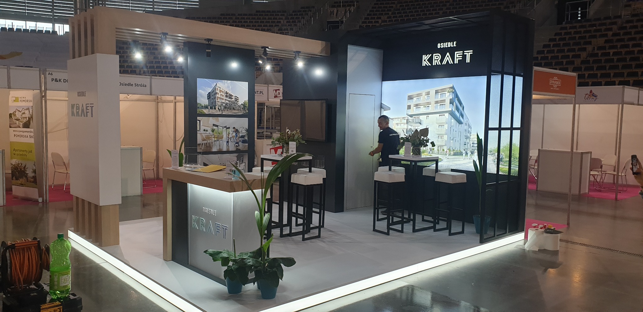 Stoisko targowe z logo Osiedle KRAFT, prezentujące wizualizacje budynków mieszkalnych, z wysokimi stołkami barowymi i roślinami doniczkowymi jako elementami dekoracyjnymi.
