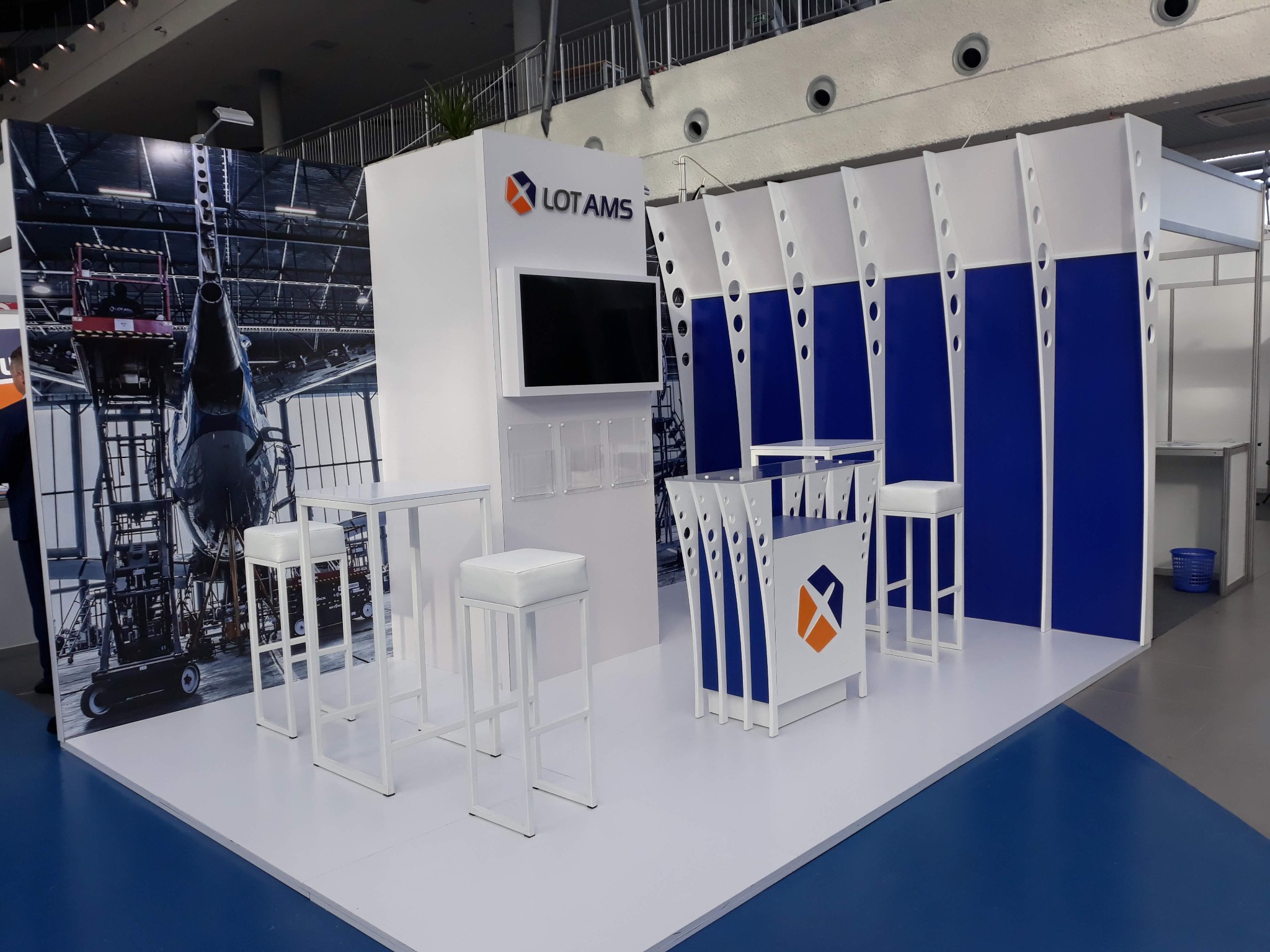 Biało-niebieskie stoisko targowe LOT AMS z logo, ekranem i grafiką samolotu w tle, wyposażone w białe stoliki i hokery.