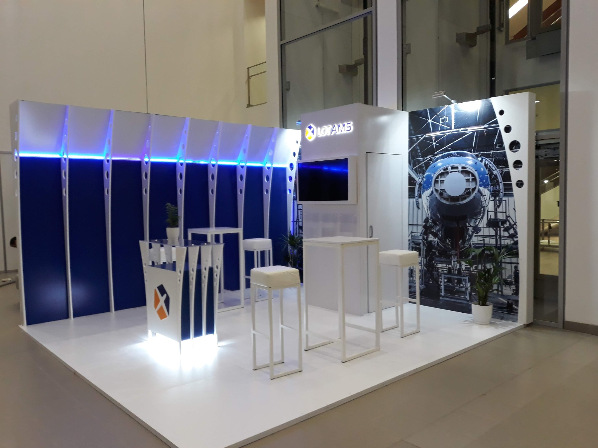 Nowoczesne stoisko targowe z niebieskimi akcentami i podświetleniem LED, wyposażone w białe stoliki barowe i hokery, z grafiką przedstawiającą złożoną maszynę na ścianie.