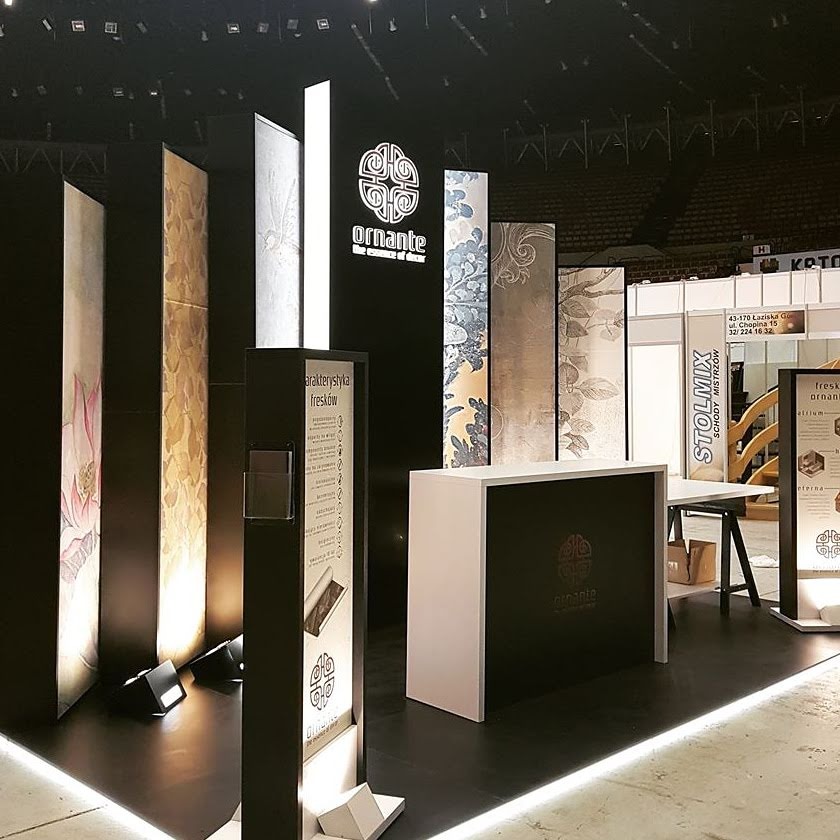 Ekspozycja paneli dekoracyjnych z motywami roślinnymi na stoisku targowym, oświetlenie punktowe, logo firmy Ornante, charakterystyka fresków na panelu informacyjnym.