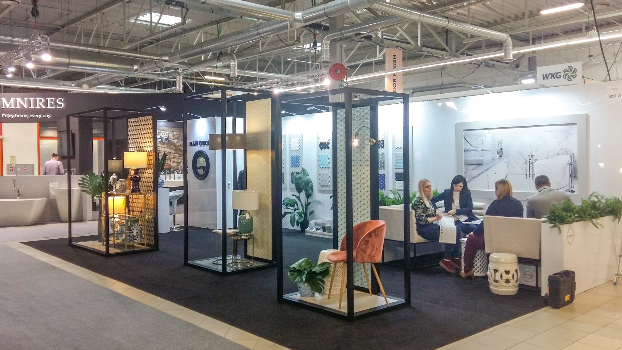 Aranżacja stoiska targowego z ekspozycją łazienek i elementów wyposażenia wnętrz, w tym umywalki, armatura, oświetlenie i płytki ceramiczne, z widocznymi osobami w tle.