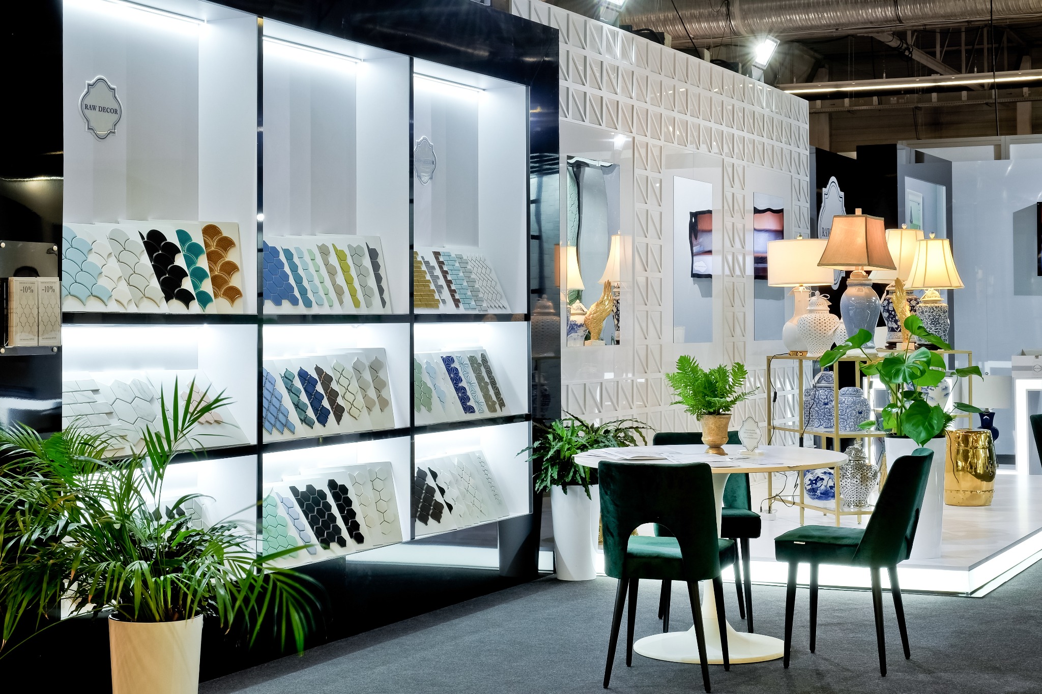 Ekspozycja płytek mozaikowych w kształcie rybich łusek i heksagonów w różnych kolorach, na tle białych regałów z oświetleniem, z elementami aranżacji wnętrz: zielone krzesła, okrągły stolik...