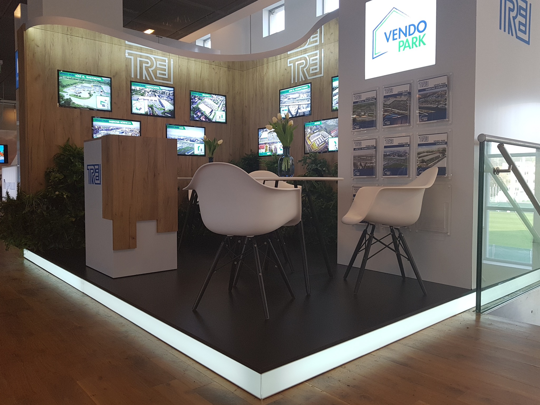 Stoisko targowe firmy TRE z drewnianą zabudową, białymi krzesłami i monitorami prezentującymi parki handlowe Vendo Park, oświetlone ledami przy podłodze.