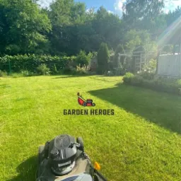 Kosiarka do trawy na tle wypielęgnowanego trawnika z widocznym ogrodem i drzewami w słoneczny dzień. Na trawniku widoczny znak wodny 'Garden Heroes'.