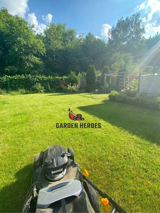 Kosiarka do trawy na tle wypielęgnowanego trawnika z widocznym ogrodem i drzewami w słoneczny dzień. Na trawniku widoczny znak wodny 'Garden Heroes'.