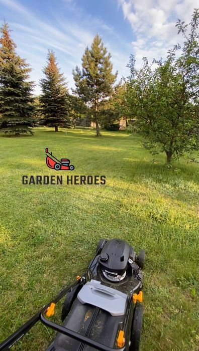 Elektryczna kosiarka do trawy na tle zielonego trawnika i drzew iglastych w słoneczny dzień. Na trawniku widoczny znak graficzny i napis 'GARDEN HEROES'.