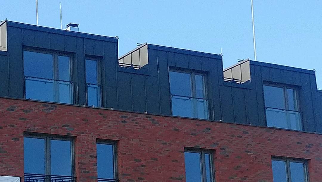 Fragment fasady budynku mieszkalno-usługowego z czerwonej cegły, z widocznymi oknami i balkonami z przeszklonymi balustradami, zwieńczonego ciemną blachą na dachu.