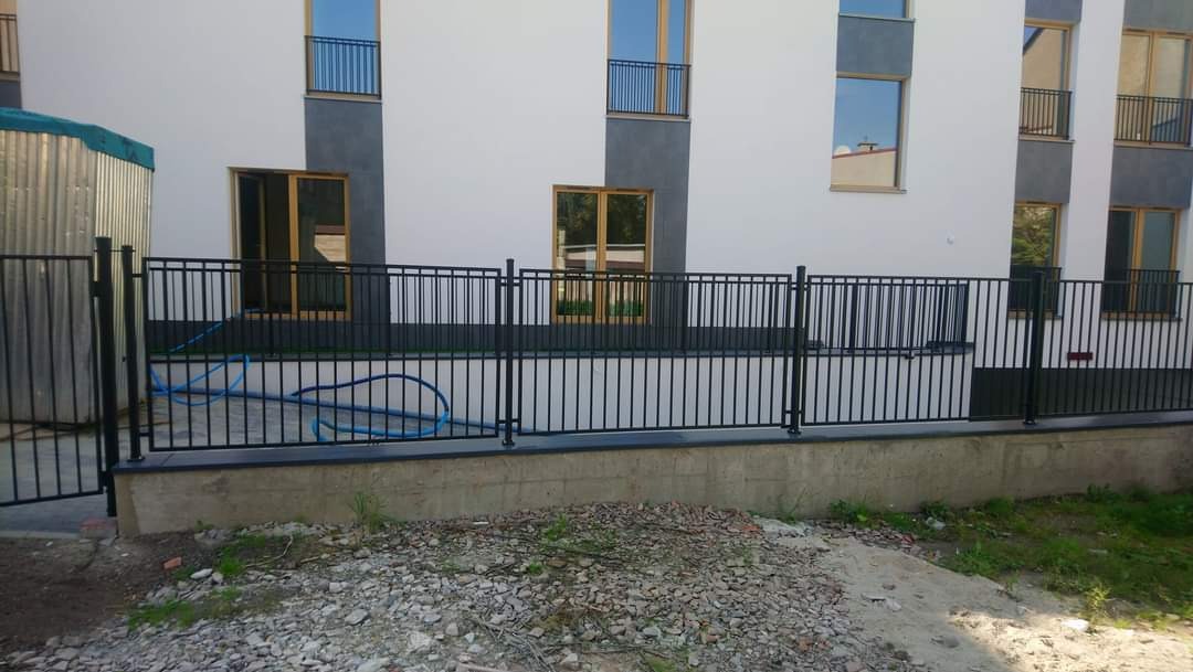 Czarna metalowa barierka zamontowana przed budynkiem mieszkalnym z widocznym niebieskim wężem ogrodowym. Barierka posiada pionowe pręty i poziome elementy łączące, a jej fragment stanowi furtkę.