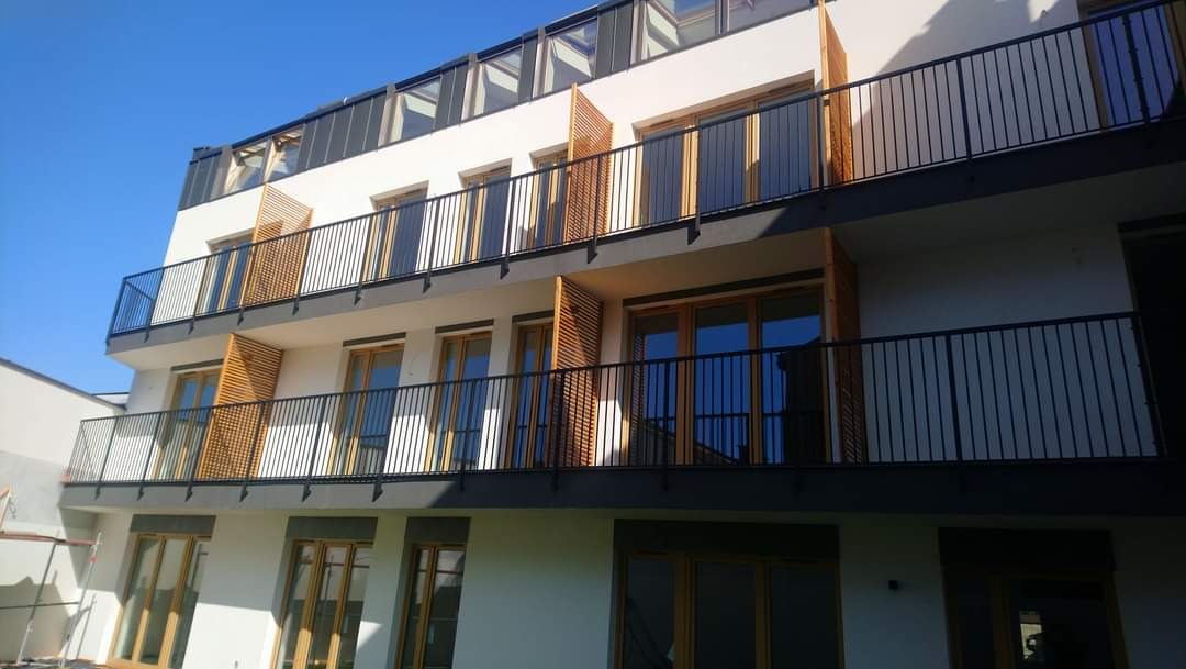 Nowoczesny budynek mieszkalny z balkonami zabezpieczonymi czarnymi, metalowymi balustradami i drewnianymi żaluzjami balkonowymi, widok z dołu na dwa piętra.
