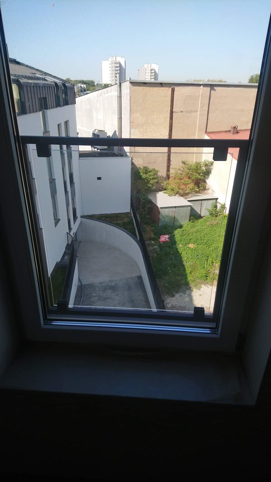 Widok z okna na szklaną balustradę balkonu, betonowy podjazd i zielony ogród z budynkami w tle, w słoneczny dzień.