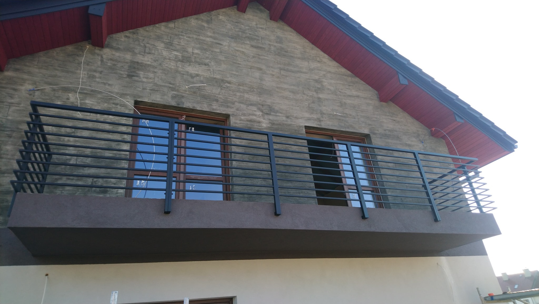 Czarna, pozioma balustrada metalowa z prostymi prętami, zamontowana na balkonie domu z elewacją imitującą drewno, widoczna z dołu pod kątem.