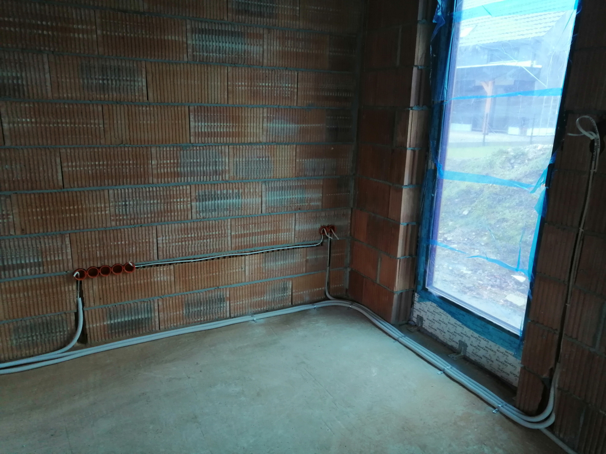 Wnętrze w trakcie budowy z widoczną instalacją elektryczną: kable w szarych rurkach prowadzone wzdłuż ściany z cegieł, zakończone pomarańczowymi puszkami pod gniazdka, okno zabezpieczone folią w tle.