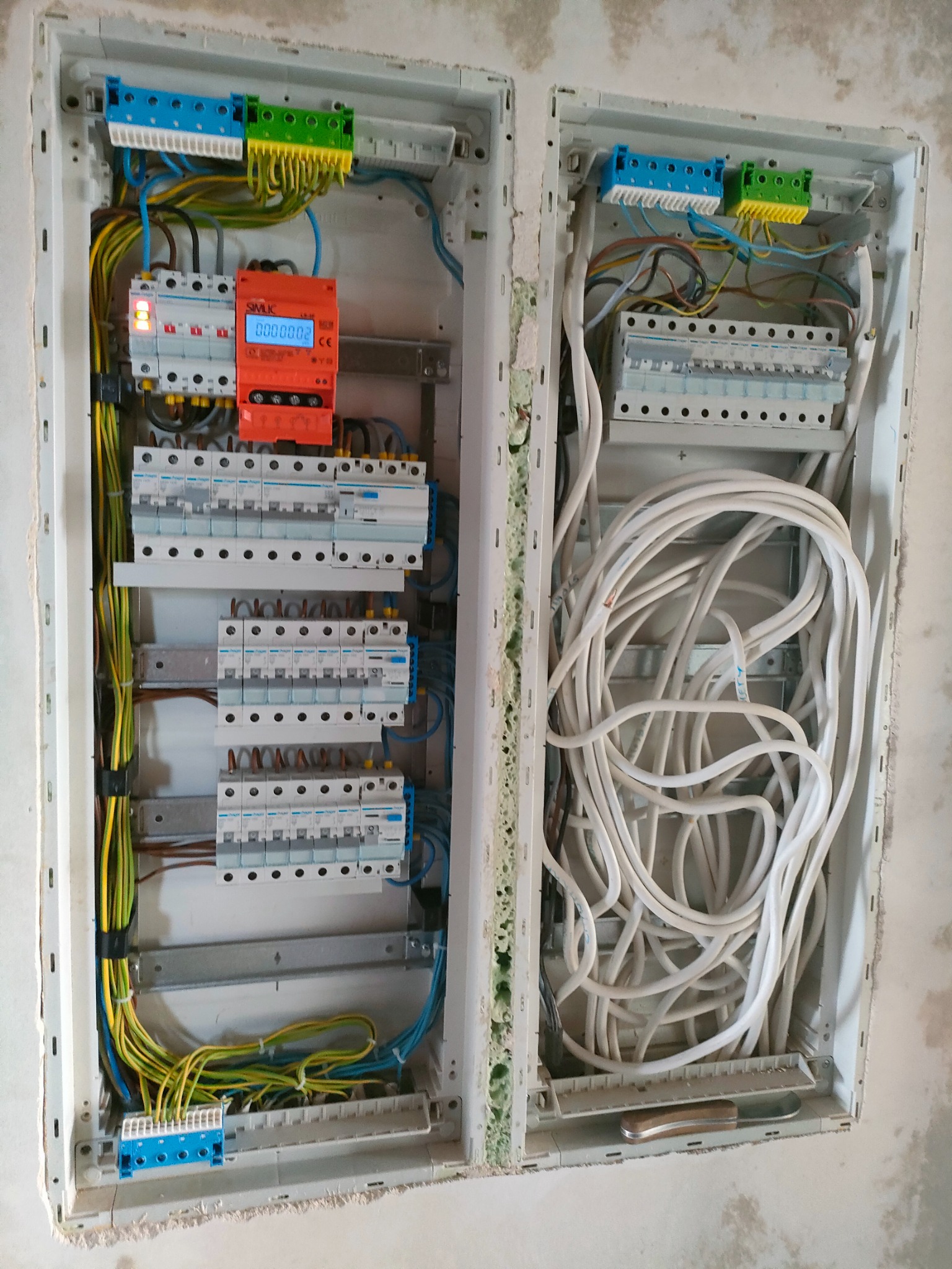 Dwa panele elektryczne w trakcie montażu, jeden z zamontowanymi bezpiecznikami i miernikiem zużycia energii, drugi z nieułożonymi kablami i scyzorykiem na dole.