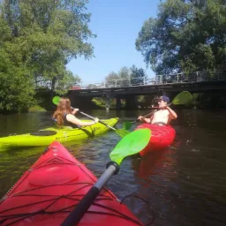 Trzy kajaki na rzece, z widocznym mostem w tle i osobami wiosłującymi, jeden kajak z wiosłem skierowany w stronę widza.