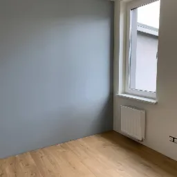 Pomalowany na szaro pok&oacute;j z drewnianą podłogą, białym oknem i grzejnikiem pod nim, widoczne gniazdka elektryczne.