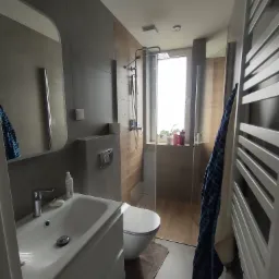 Nowoczesna łazienka z szarą armaturą, kabiną prysznicową ze szklaną ścianą, umywalką z szafką, toaletą i grzejnikiem drabinkowym z powieszonym niebieskim ręcznikiem.