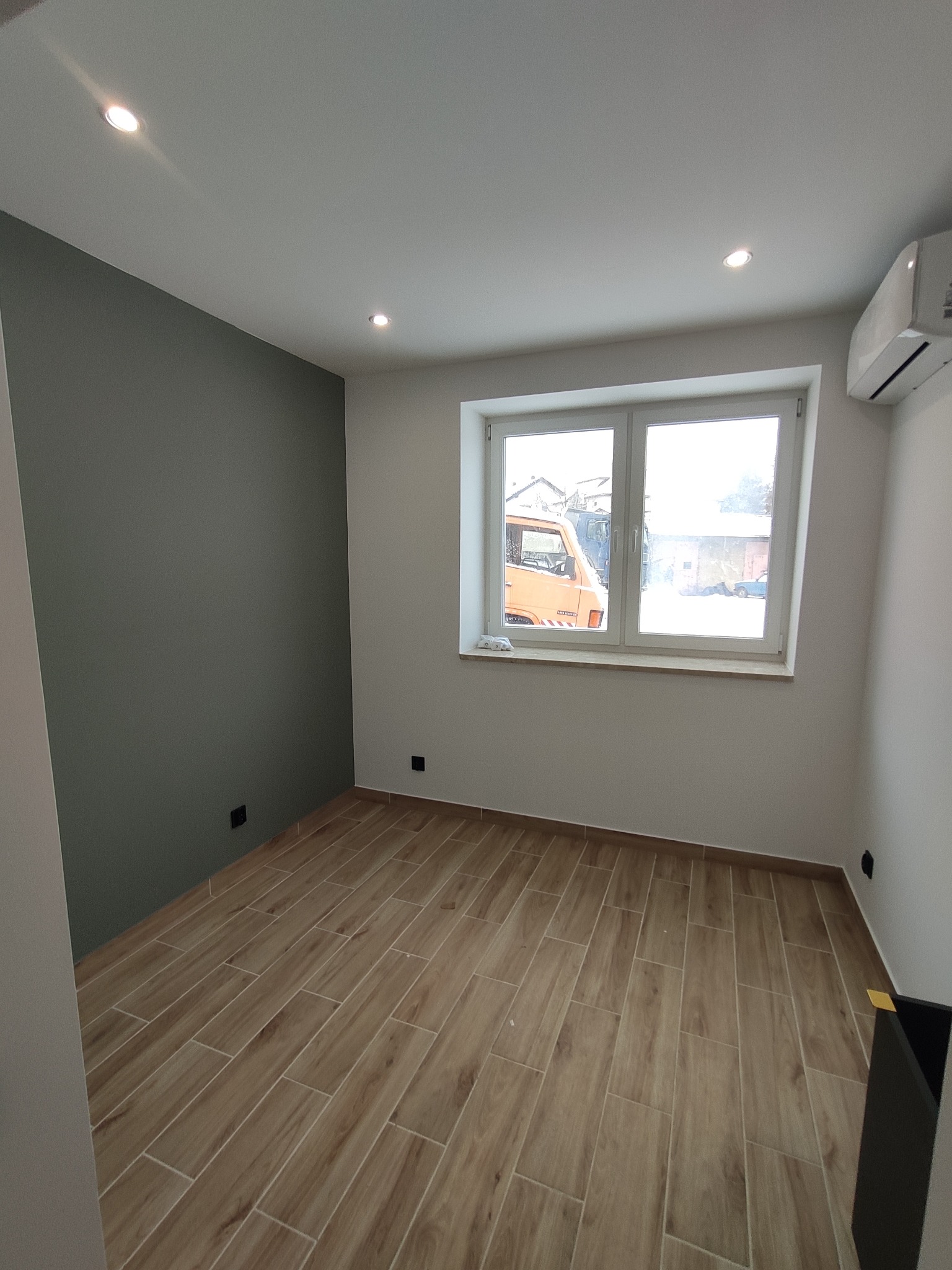 Wnętrze pokoju po remoncie: pomalowana szara ściana, ułożona podłoga z paneli imitujących drewno, białe ściany, okno z widokiem na pomarańczowy samochód dostawczy, zamontowany klimatyzator.