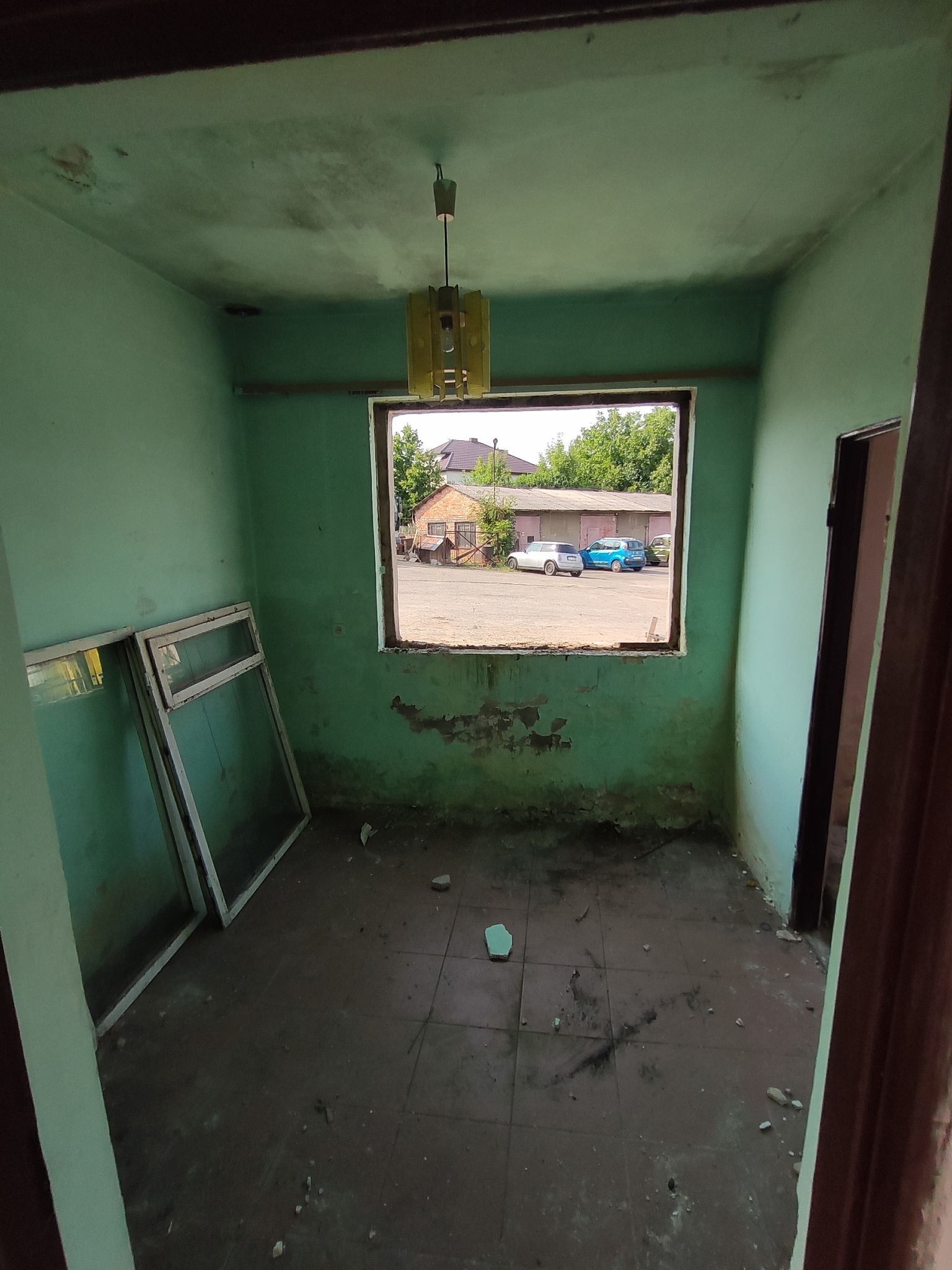 Wnętrze pokoju z zielonymi ścianami w stanie przed remontem: widoczne stare okna oparte o ścianę, okno bez szyby z widokiem na parking z samochodami, stara lampa wisząca na suficie, zniszczona...