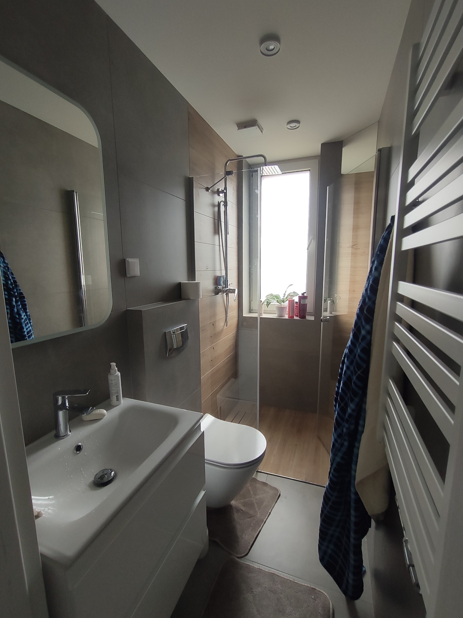 Nowoczesna łazienka z szarą armaturą, kabiną prysznicową ze szklaną ścianą, umywalką z szafką, toaletą i grzejnikiem drabinkowym z powieszonym niebieskim ręcznikiem.