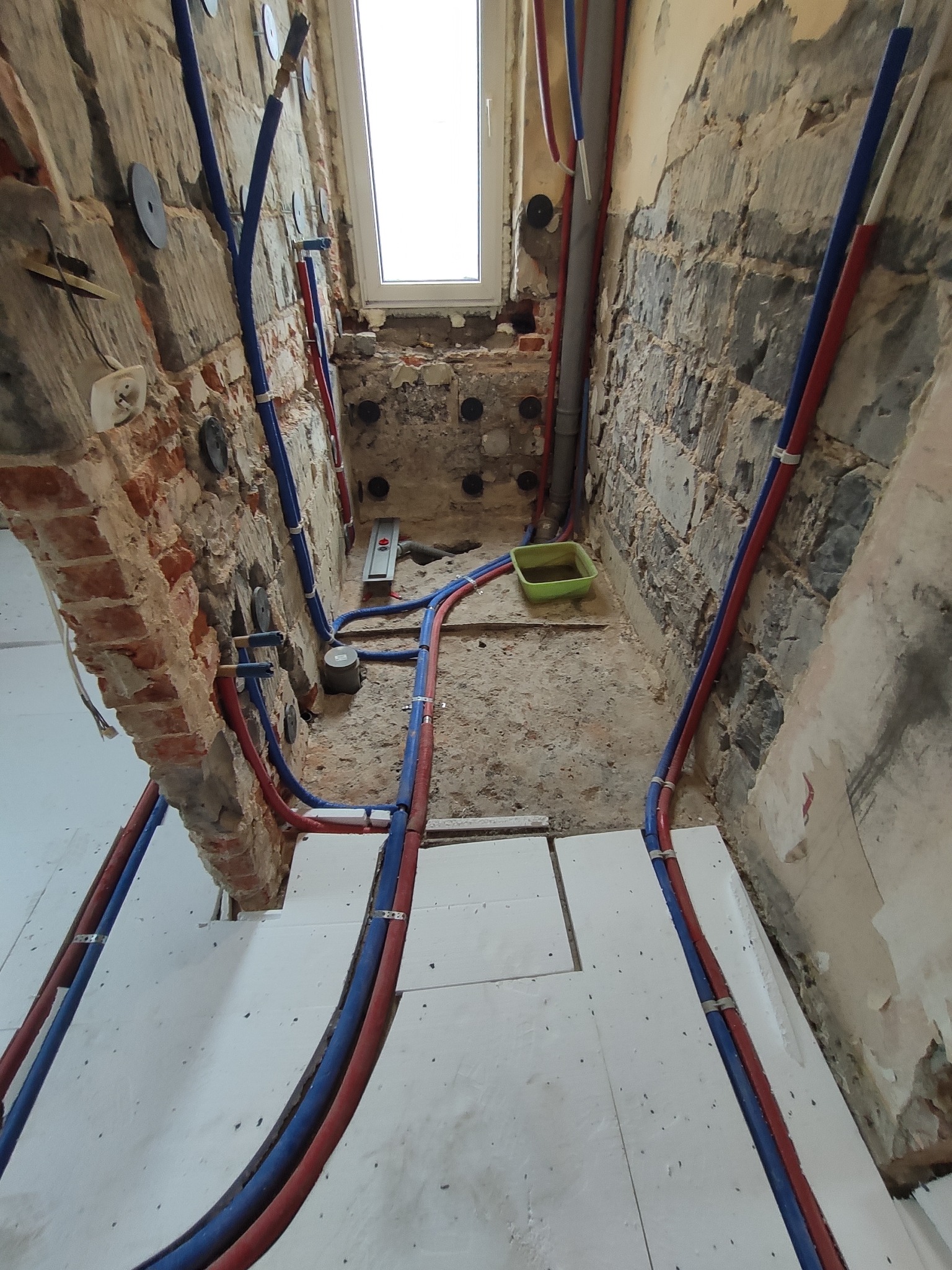 Wnętrze w trakcie remontu z odsłoniętą cegłą, nową instalacją wodną z rurami w kolorze niebieskim i czerwonym, przygotowane podłoże pod posadzkę i małe okno w głębi.