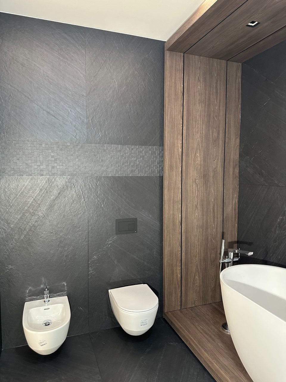 Nowoczesna łazienka z czarną kamienną okładziną ścian, mozaikowym pasem dekoracyjnym, wiszącą toaletą i bidetem oraz fragmentem wolnostojącej białej wanny z chromowaną armaturą i drewnianą zabudową.