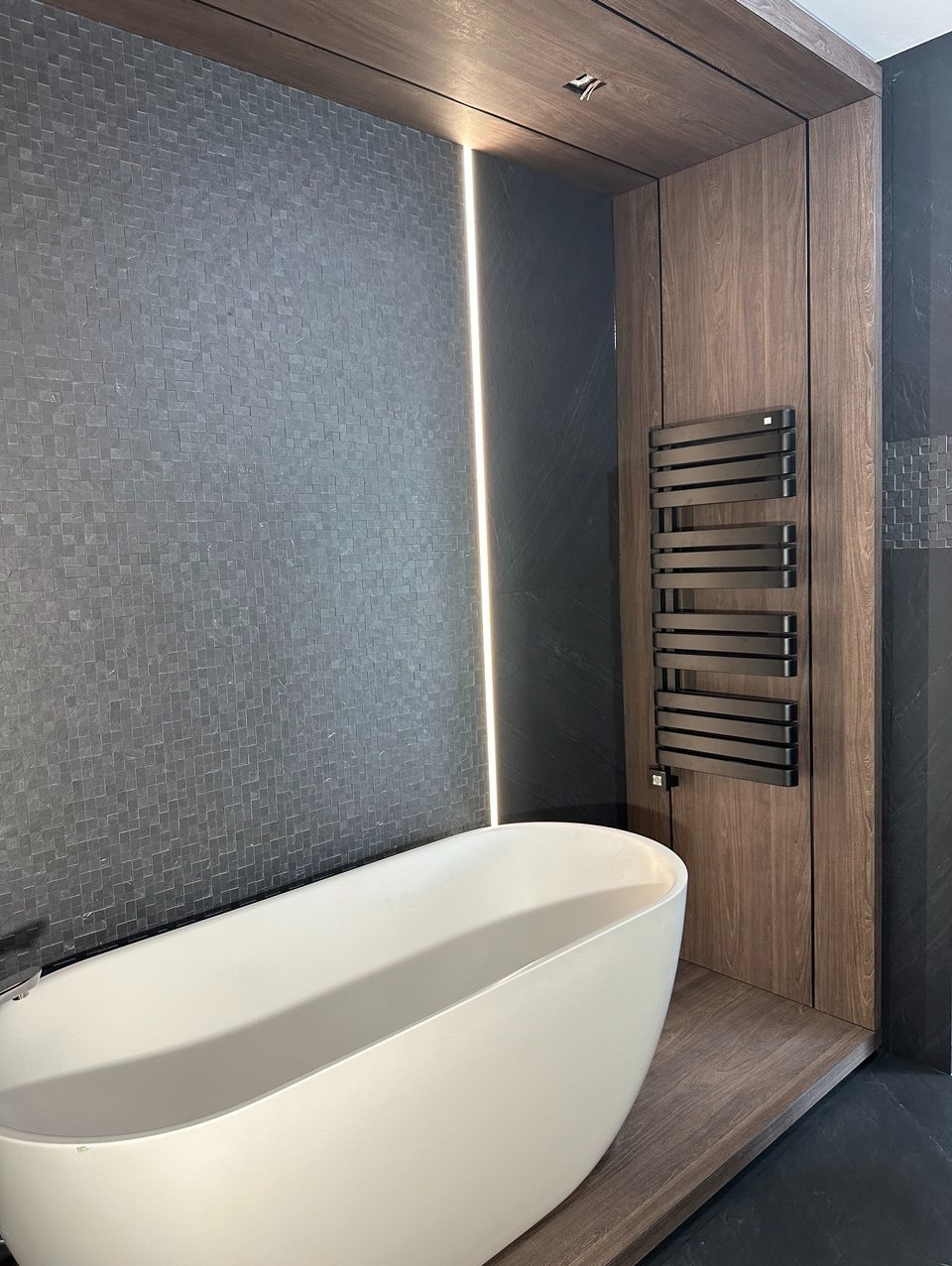 Nowoczesna łazienka z wolnostojącą białą wanną, ścianą wyłożoną drobną, szarą mozaiką, podświetleniem LED i grzejnikiem drabinkowym na tle drewnianej zabudowy.