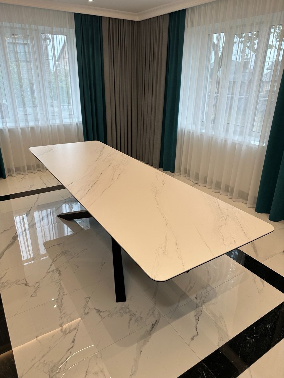 Długi stół z blatem imitującym marmur, wsparty na czarnej, pojedynczej nodze, na tle marmurowej podłogi i okien z zasłonami w odcieniach zieleni i szarości.