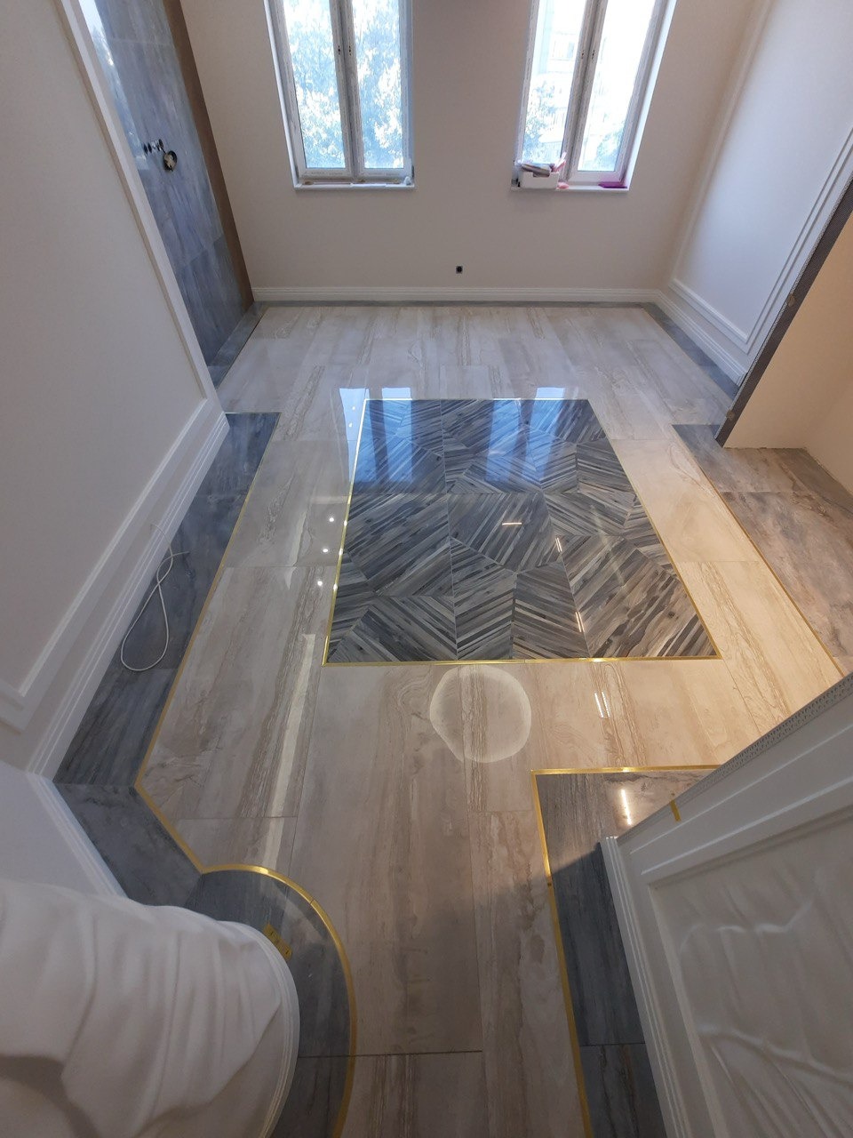 Ukończona podłoga z płytek w odcieniach beżu i szarości z dekoracyjnymi złotymi listwami, ułożona we wzór kwadratu z centralnym motywem jodełki, widok z góry.