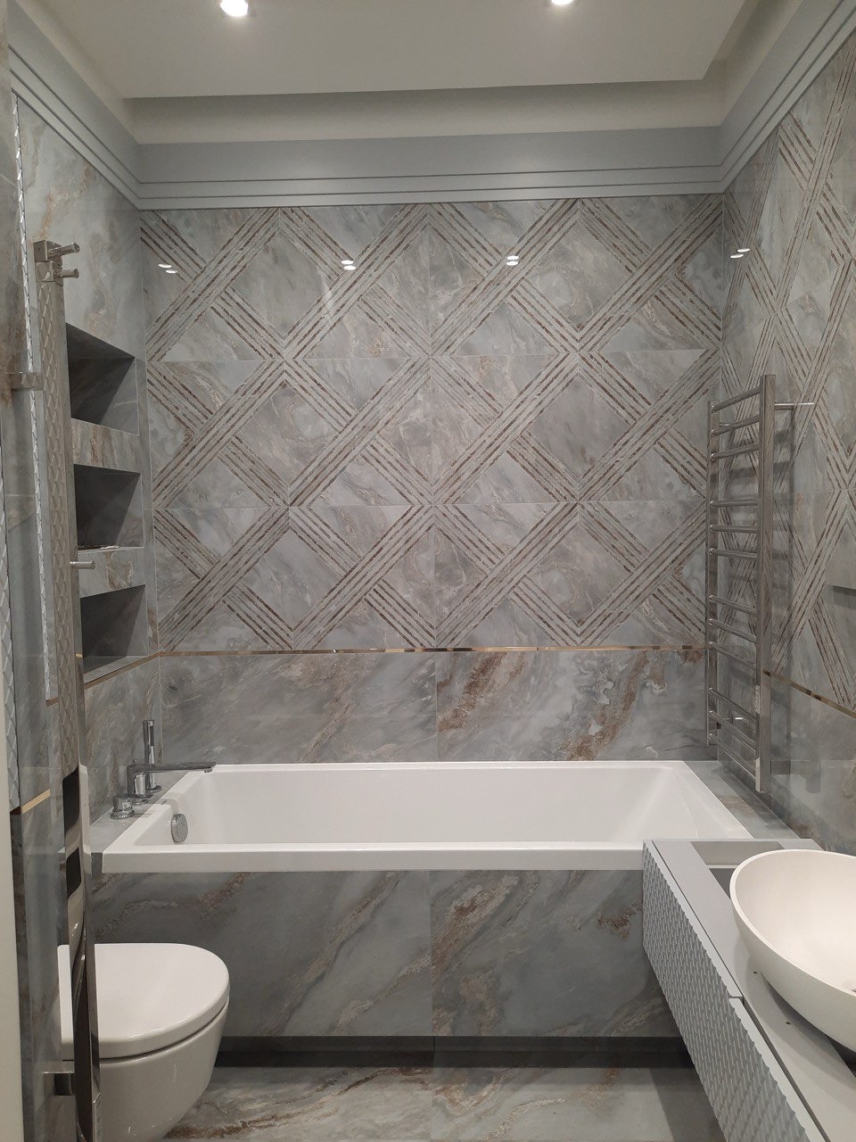Nowoczesna łazienka z marmurowymi płytkami w odcieniach szarości i beżu, biała wanna zabudowana płytkami, grzejnik drabinkowy i toaleta.