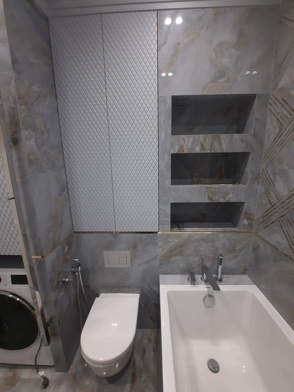 Nowoczesna łazienka z marmurowymi płytkami, zabudową nad toaletą i wanną oraz wnękami w ścianie.