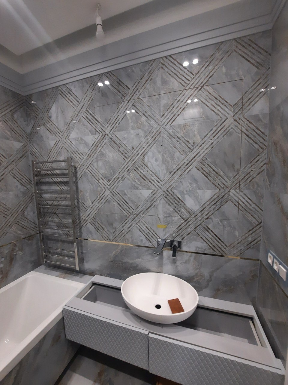 Wykończona łazienka z marmurowymi płytkami w geometryczne wzory, owalną umywalką na szarej szafce i grzejnikiem drabinkowym.