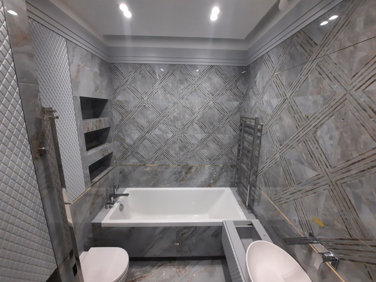 Nowoczesna łazienka w odcieniach szarości z marmurkowymi płytkami, geometrycznym wzorem na ścianach, białą wanną i umywalką, oraz grzejnikiem drabinkowym.