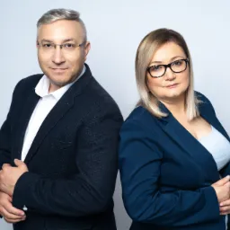 Profesjonalny portret studyjny: mężczyzna w garniturze i okularach oraz kobieta w niebieskiej marynarce, stojący plecami do siebie na jasnym tle.