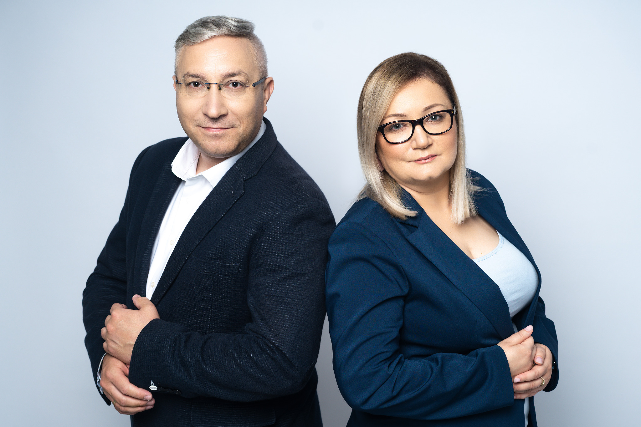 Profesjonalny portret studyjny: mężczyzna w garniturze i okularach oraz kobieta w niebieskiej marynarce, stojący plecami do siebie na jasnym tle.