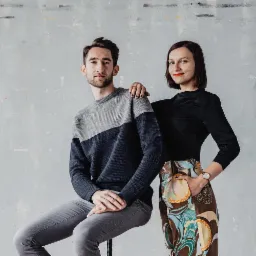 Młody mężczyzna w dwukolorowym swetrze siedzi na stołku, obok stoi kobieta w czarnej bluzce i wzorzystych spodniach, opierając dłoń na jego ramieniu, na tle szarej ściany.