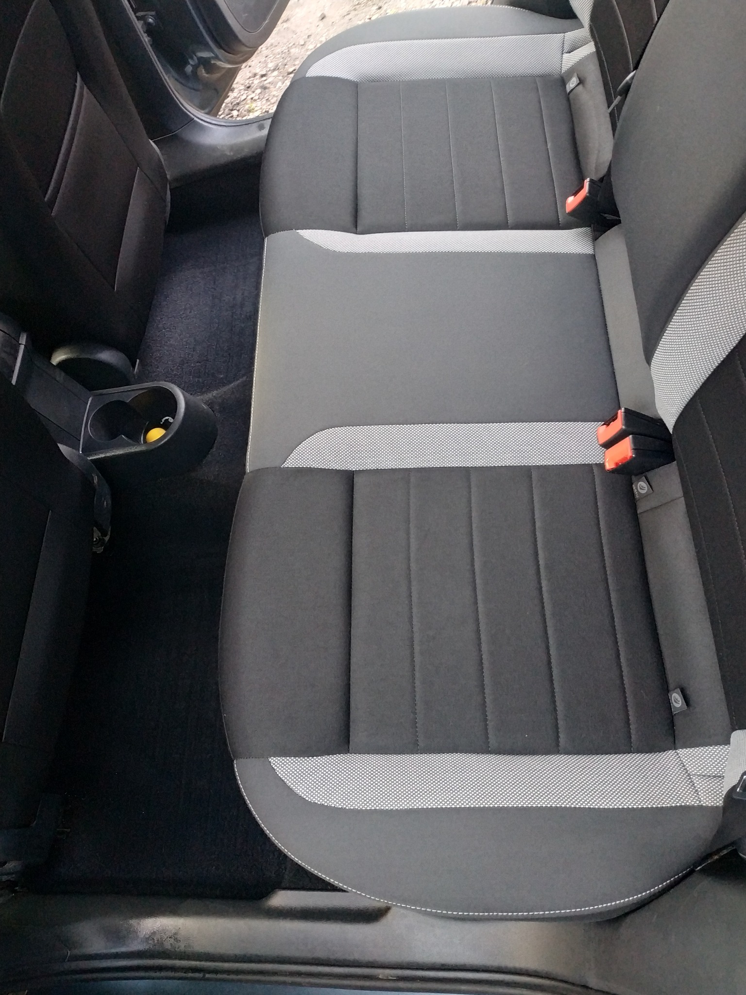 Wnętrze samochodu z widoczną tylną kanapą w odcieniach szarości, z detalami w postaci pasów bezpieczeństwa i uchwytu na napoje z żółtym elementem w środku.