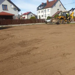 Żółta koparko-ładowarka na wyrównanym, brązowym terenie w słoneczny dzień, w tle zabudowa jednorodzinna i drewniany płot.