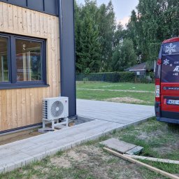 AC System Tomasz Urbański - Zamontowany klimatyzator Gree obok drewnianego domu, z tyłu czerwony bus firmy A/C System z certyfikatem montażu klimatyzacji i numerem telefonu.
