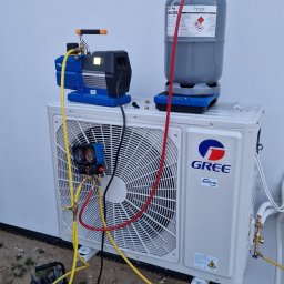 AC System Tomasz Urbański - Biała jednostka zewnętrzna klimatyzacji Gree z podłączonymi narzędziami serwisowymi: manometrami, pompą próżniową i butlą z czynnikiem chłodniczym R32. Widoczne przewody i złączki.