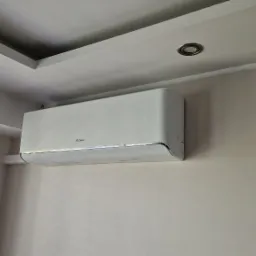 Biały klimatyzator Gree zamontowany na ścianie w pomieszczeniu, widoczne drewniane żaluzje i sufit z oświetleniem punktowym.