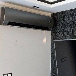 AC System Tomasz Urbański - Nowoczesny, grafitowy klimatyzator ścienny zamontowany w pomieszczeniu z wzorzystą tapetą i ciemną zasłoną. Widoczny wyświetlacz temperatury.