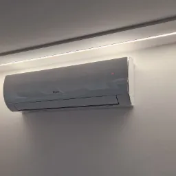 Zamontowany klimatyzator Gree na białej ścianie, oświetlony listwą LED przy suficie, z fragmentem okna z żaluzjami w tle. Nowoczesny design i minimalistyczny styl.