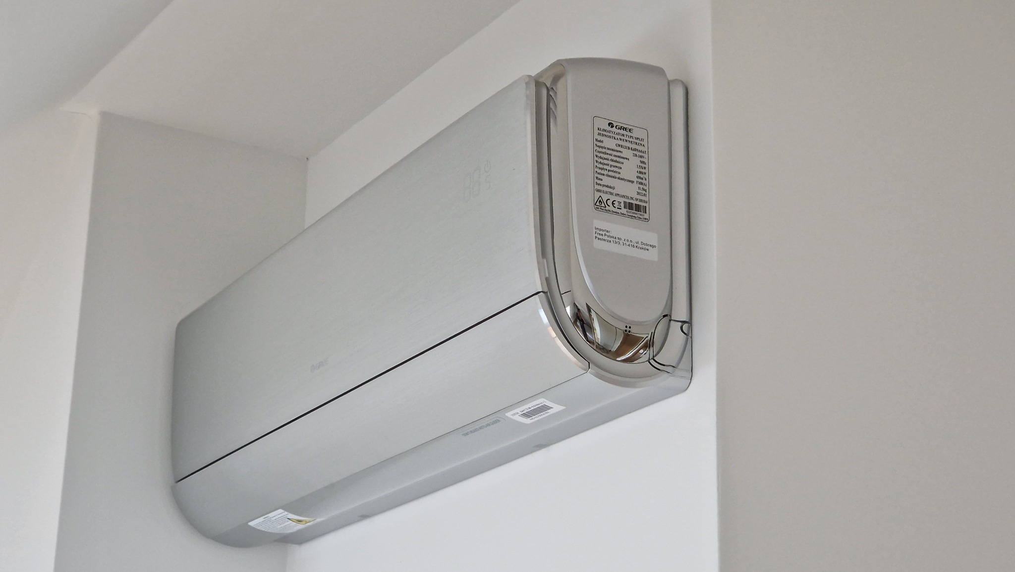 Biały klimatyzator marki Gree zamontowany na białej ścianie, widoczny wyświetlacz temperatury i etykieta z danymi technicznymi.