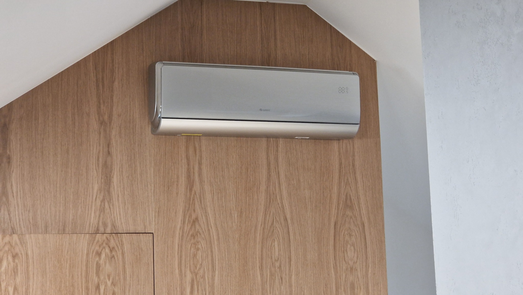 Nowoczesny, srebrny klimatyzator marki Gree zamontowany na drewnianej, skośnej ścianie poddasza.