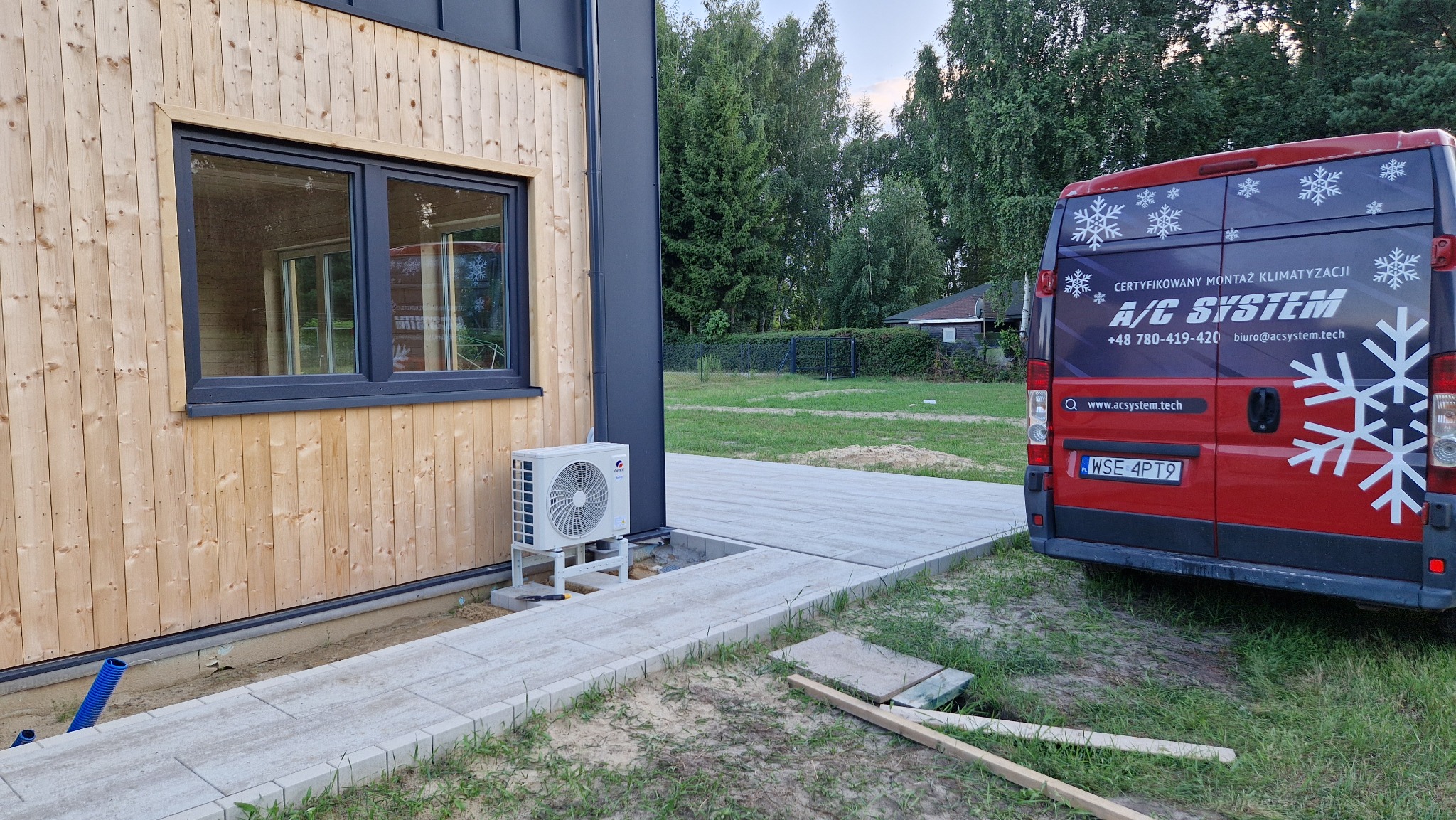 Zamontowany klimatyzator Gree obok drewnianego domu, z tyłu czerwony bus firmy A/C System z certyfikatem montażu klimatyzacji i numerem telefonu.