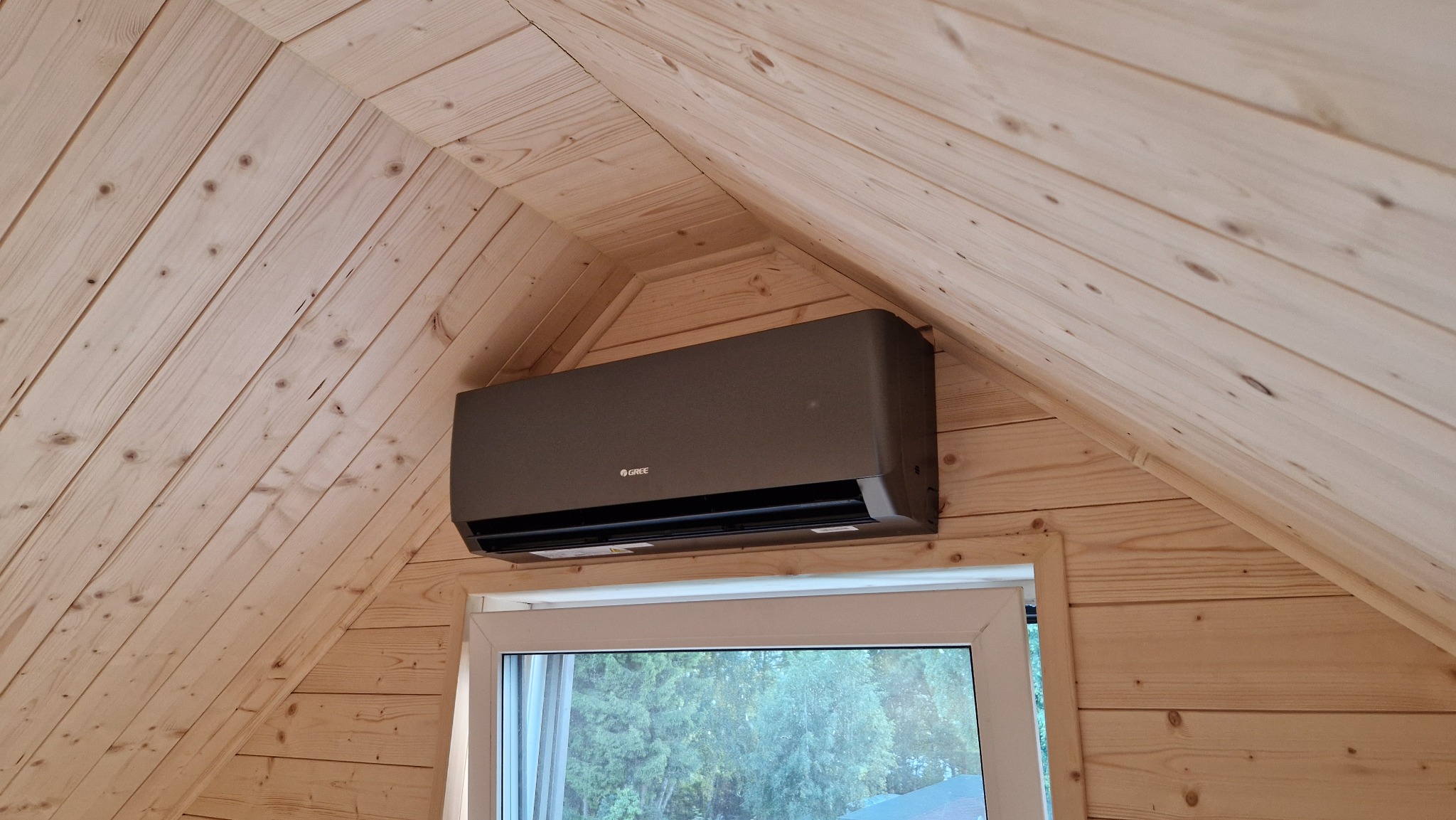 Szary klimatyzator Gree zamontowany na drewnianej ścianie pod skosem, nad oknem. Widok na zewnątrz przez okno. Naturalne światło.
