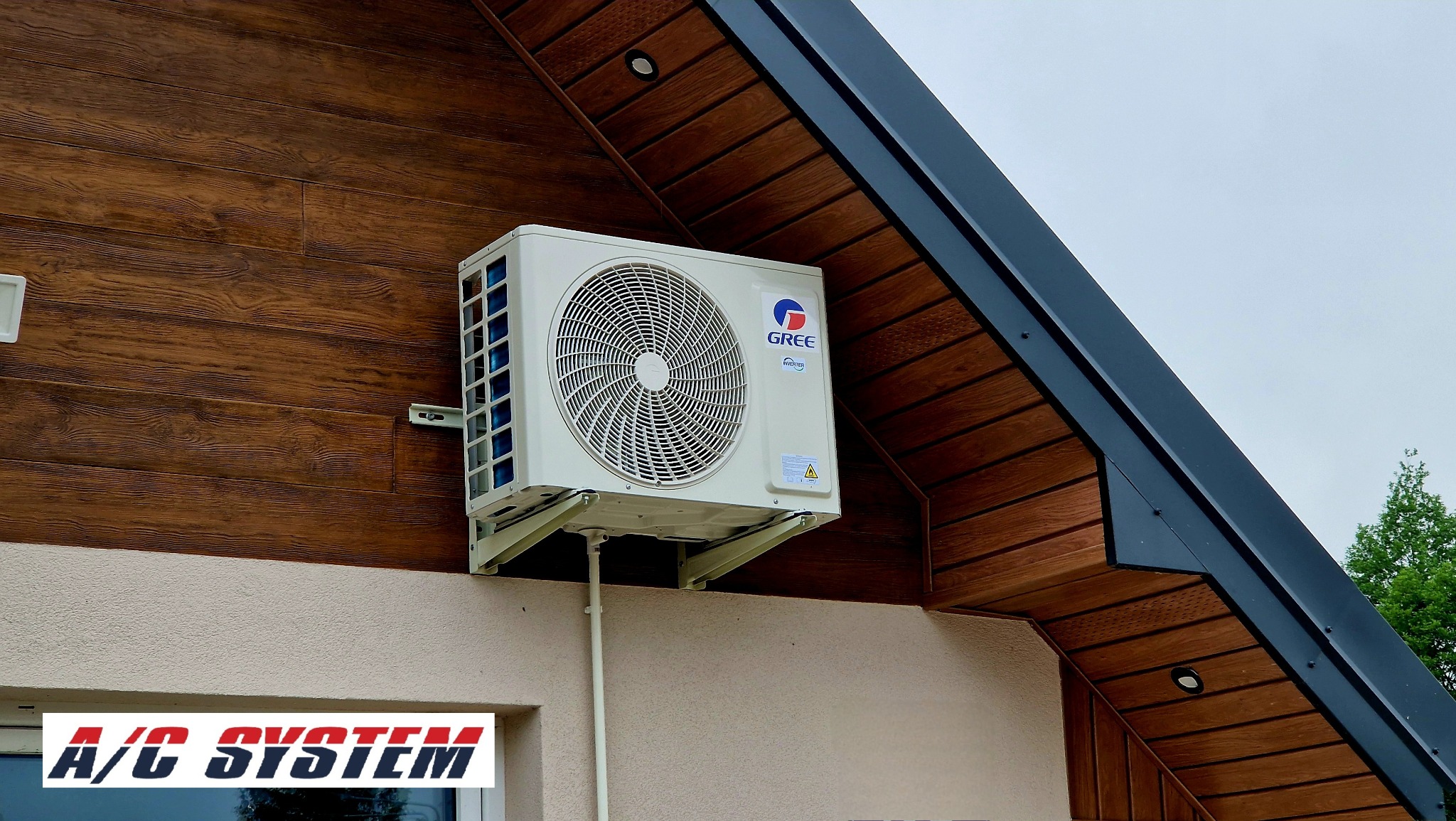 Biała jednostka zewnętrzna klimatyzacji Gree zamontowana na brązowej elewacji domu z elementami imitującymi drewno. Widoczne przewody i logo producenta.