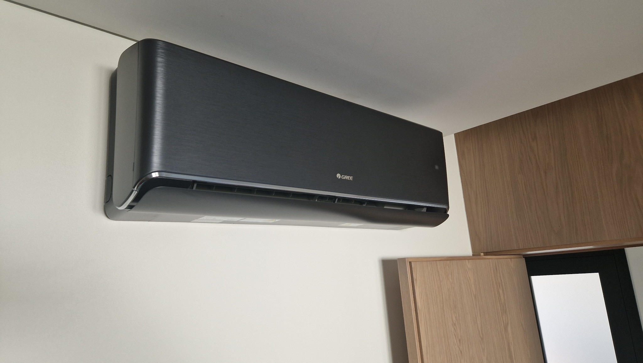 Czarny klimatyzator Gree, zamontowany na białej ścianie w pomieszczeniu z drewnianymi elementami wykończenia. Widoczny z bliska, z boku.