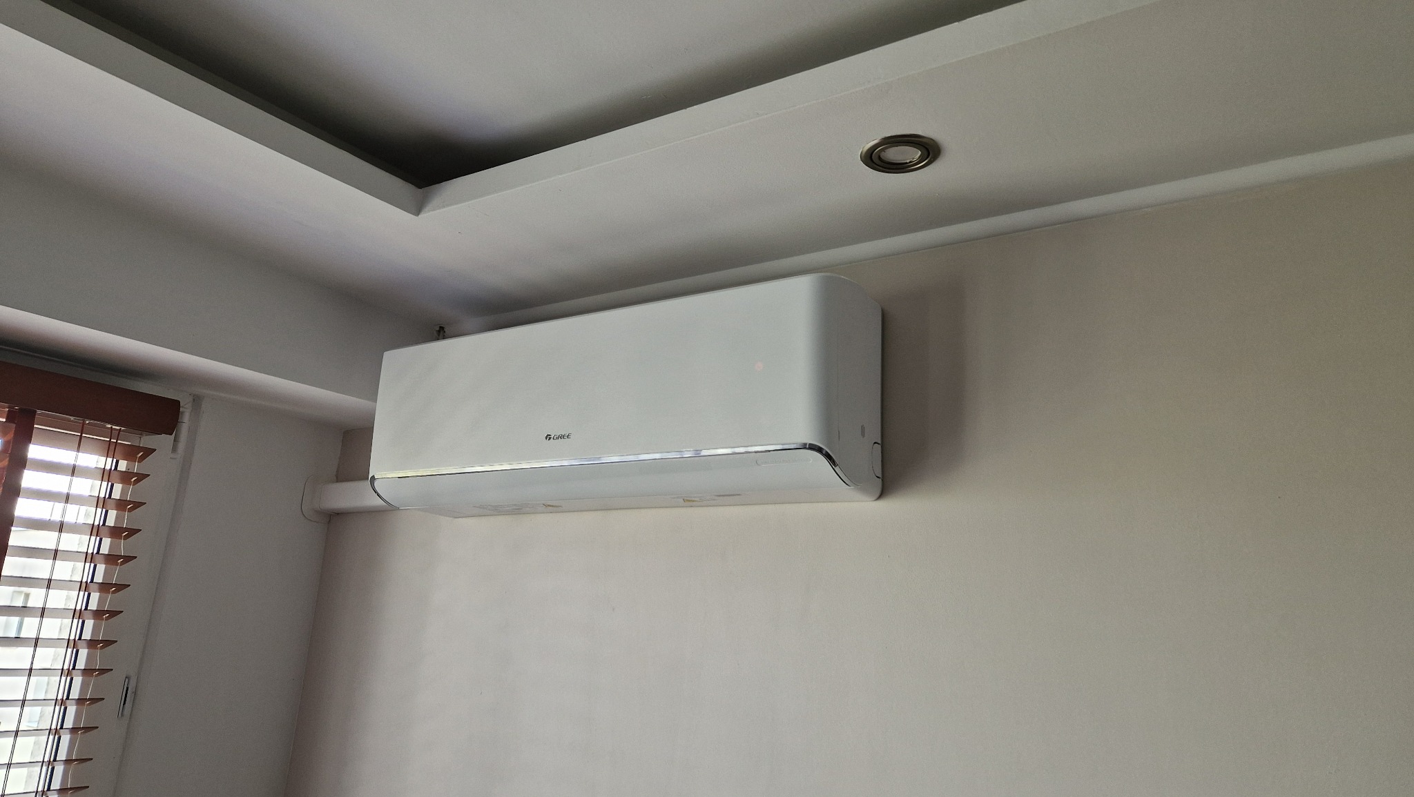 Biały klimatyzator Gree zamontowany na ścianie w pomieszczeniu, widoczne drewniane żaluzje i sufit z oświetleniem punktowym.